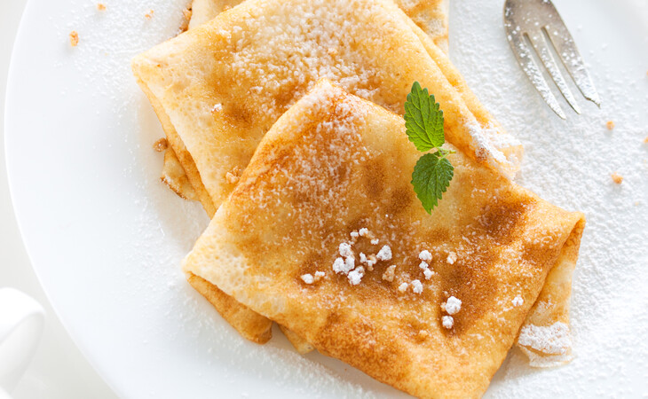 Das Bild zeigt einen weißen Teller mit Crêpes, die mit Puderzucker bestreut sind, daneben liegt eine Gabel. Die Crêpes wirken frisch zubereitet, haben eine goldbraune Farbe und sind leicht mit Puderzucker bedeckt