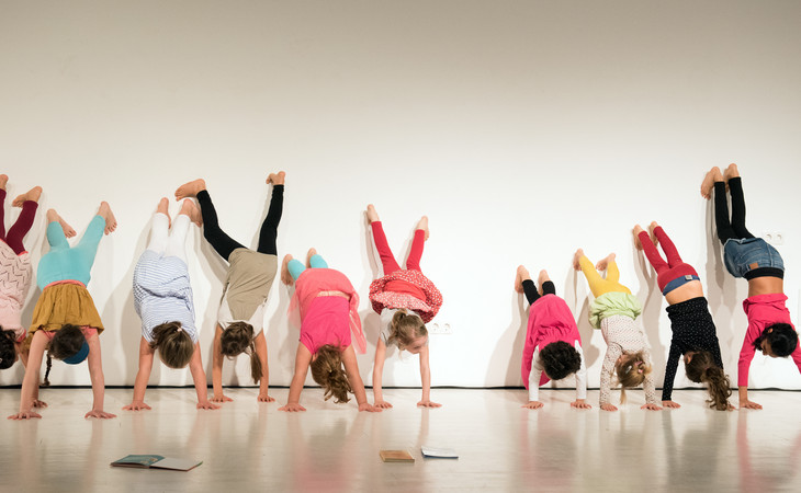 Zu sehen ist ein querformatiges Bild, auf dem Kinder einen Handstand machen. Es sind insgesamt zehn Kinder in bunter Kleidung vor einer weißen Wand. Sie sind unterschiedlich groß und stützen ihre Beine an der Wand ab, um kopfüber auf ihren Händen zu stehen. Der Boden ist hellgrau und spiegelnd, es ist die Spiegelung der Kinder darauf zu erkennen. Es liegen auch vier Bücher auf dem Boden, eines ist aufgeschlagen.