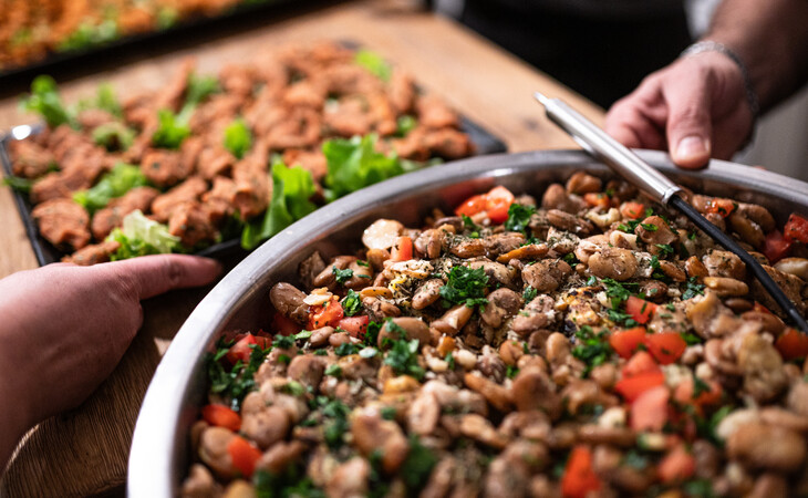 Eine Person mischt einen großen Salat in einer Schüssel auf einem Tisch, umgeben von weiteren Schalen mit vorbereitetem Essen.