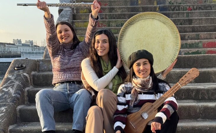 Drei junge Frauen sitzen auf einer Treppe, eine hält eine Flöte, eine eine Rahmentrommel, die dritte eine Ukulele, alle lächeln
