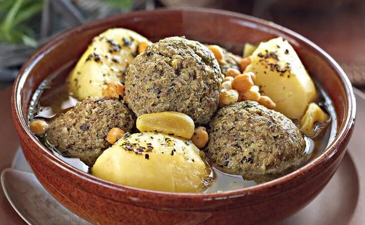 „Eine Schüssel mit Essen, vermutlich Fleischbällchen und Kartoffeln. Die Fleischbällchen sind rund und gewürzt, die Kartoffeln in kleine Würfel geschnitten und daraufgelegt. Serviert wird das Gericht in einer braunen Keramikschüssel, ein Löffel liegt daneben. Der Hintergrund ist unscharf, die Schüssel steht vermutlich auf einem Holztisch mit einer Pflanze in der Ecke. Die Farben wirken warm und einladend.