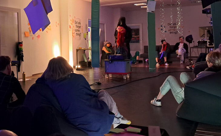 Eine Gruppe von Menschen sitzt in einem Raum mit einer Bühne in der Mitte. Die Bühne ist mit bunten Lichtern dekoriert, mehrere Personen stehen darauf. Auf der linken Seite der Bühne steht eine Person in rotem Jackett mit Mikrofon. Im Hintergrund sitzen andere Menschen auf Stühlen, einige stehen. Der Raum wirkt wie eine Galerie oder ein Ausstellungsraum mit hoher Decke und großen Fenstern. Das Publikum schaut zur Bühne und wirkt aufmerksam.