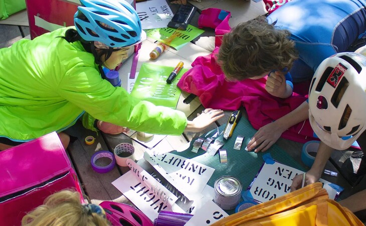 Menschen mit Fahrradhelmen sitzen um einen Tisch und gestalten mit Schablonen und Farbe Plakate
