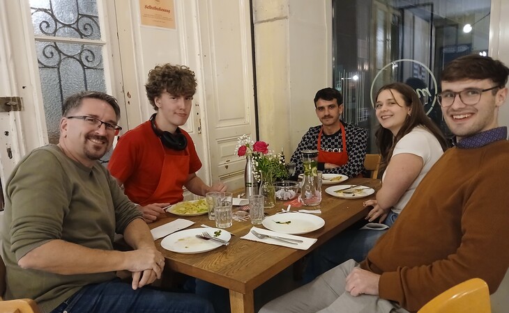 eine Gruppe von sechs Personen, die an einem Holztisch in einem Restaurant sitzen. Alle lächeln und scheinen die gemeinsame Zeit zu genießen. Der Tisch ist mit Tellern, Gläsern und einer Blumenvase gedeckt. Das Restaurant hat weiße Wände und ein großes Fenster mit Blick nach draußen. An der Wand befindet sich ein Schild mit der Aufschrift „Restaurant“. Die Personen tragen legere Kleidung und sitzen auf Stühlen rund um den Tisch.