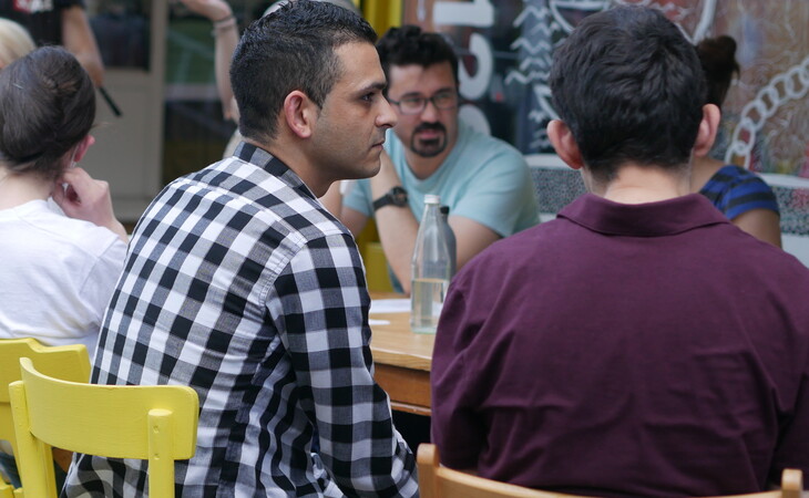 Eine Gruppe von Menschen sitzt an einem Tisch draußen vor einem Restaurant. Auf dem Bild sind sechs Personen zu sehen – drei Männer und drei Frauen – alle tragen Gesichtsmasken. Der Mann in der Mitte trägt ein schwarz-weiß kariertes Hemd und scheint mit den beiden anderen Männern zu sprechen. Der Tisch ist mit einer weißen Tischdecke bedeckt und um ihn herum stehen gelbe Stühle. Das Restaurant hat ein großes Fenster mit Graffiti darauf und ein Schild mit der Aufschrift ‚Cafe‘. In der Umgebung laufen weitere Menschen herum und einige sitzen auf Hockern. Die allgemeine Atmosphäre des Bildes wirkt locker und entspannt.