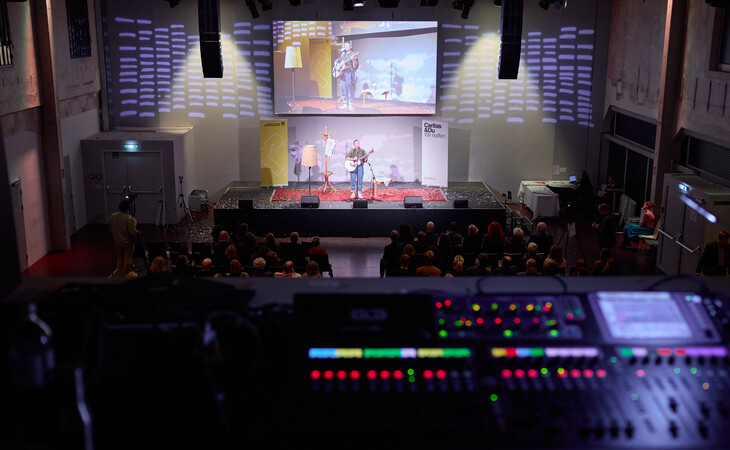 Blick von der Tonregie auf eine Bühne mit zwei Musikern und ein Publikum in einem Veranstaltungsraum mit Lichtprojektionen an der Wand