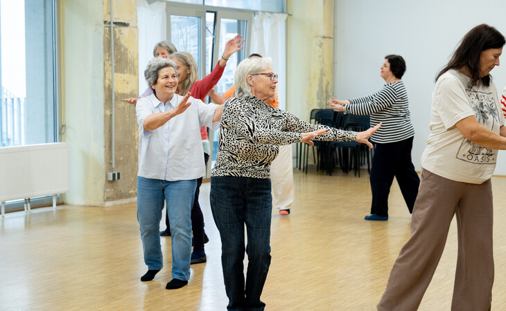Eine Gruppe älterer Menschen nimmt an einem Tanzkurs teil. Sie befinden sich in einem großen Raum mit Holzboden und großen Fenstern im Hintergrund. Die Menschen stehen in einer Reihe, mit zur Seite ausgestreckten Armen und an den Knien gebeugten Beinen. Sie scheinen sich mitten in einer Tanzbewegung zu befinden, ihre Hände und Füße sind in der Luft. Einige tragen legere Kleidung, während andere etwas eleganter gekleidet sind. Die Gruppe besteht aus Menschen unterschiedlichen Alters und Geschlechts, und alle lächeln und haben Spaß.