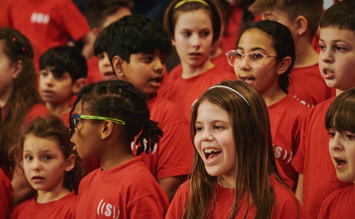 Gruppe von Kindern in roten T-Shirts singt gemeinsam, einige mit Brillen und Haarbändern