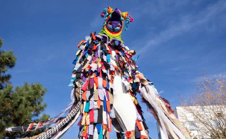 Eine Person trägt ein großes, farbenfrohes Ganzkörperkostüm aus bunten Stoffstreifen vor blauem Himmel im Freien.