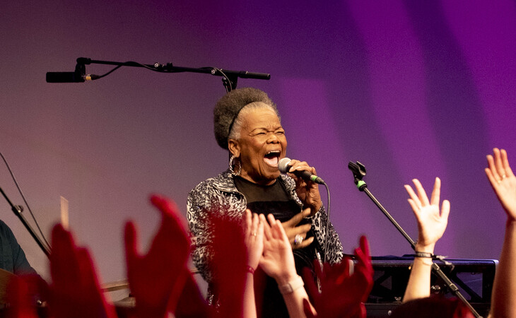 Das Bild zeigt eine ältere afroamerikanische Frau, die auf einer Bühne in ein Mikrofon singt. Sie trägt ein schwarz-weiß gemustertes Oberteil und hat ein großes Lächeln im Gesicht. Die Bühne ist mit violettem und blauem Licht beleuchtet, und vor ihr steht ein Mikrofonständer. Im Vordergrund ist das Publikum zu sehen, das die Hände in die Luft hebt, jubelt und klatscht. Das Bild wirkt, als wäre es während eines Konzerts oder einer Aufführung aufgenommen worden.
