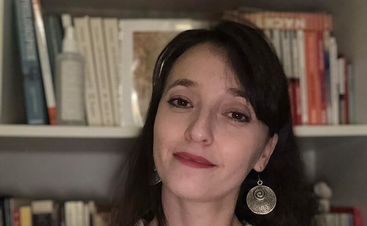 Eine Person mit langen dunklen Haaren steht vor einem Bücherregal und blickt in die Kamera.