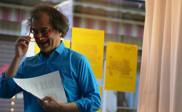 Eine Person in einem blauen Hemd steht am Fenster, liest aus einem Blatt Papier und lächelt.
