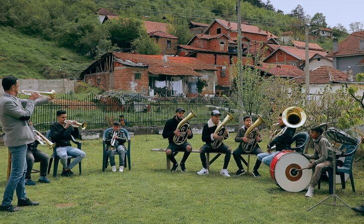 Mehrere Musiker sitzen auf Stühlen im Freien und spielen gemeinsam Blasinstrumente vor einem Dorf mit roten Dächern.