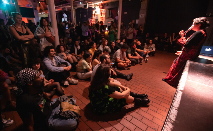 Publikum sitzt auf dem Boden und hört einer Performerin in rotem Kleid auf einer Bühne zu, die im Rahmen eines Storytelling- und Musikprogramms spricht.