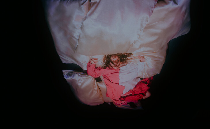 Eine Person liegt in rosafarbener Kleidung auf großen weißen Kissen, von oben fotografiert vor dunklem Hintergrund.