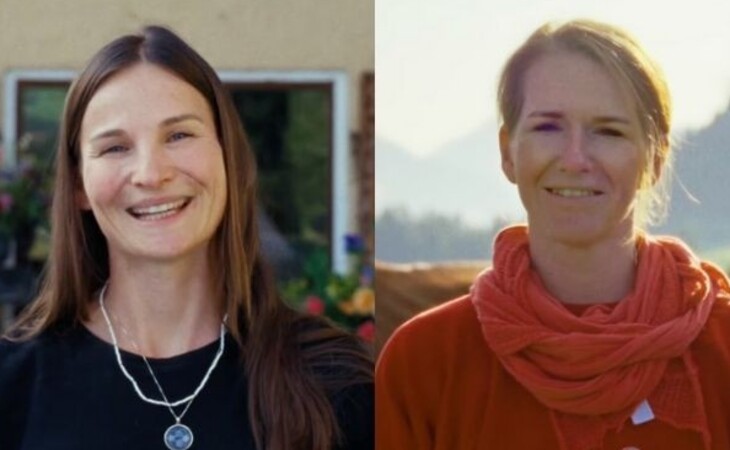 Das Bild zeigt zwei Frauen, die nebeneinander vor einem Haus stehen und beide lächeln. Die Frau links trägt ein schwarzes Kleid und eine Halskette, während die Frau in der Mitte ein orangefarbenes Kleid trägt. Der Hintergrund des Bildes ist unscharf, aber man kann ein Haus mit einem Fenster, Pflanzen, Bäumen, Bergen und dem Himmel erkennen