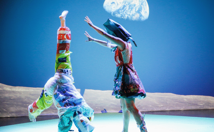 Zwei Personen mit bunten Kostümen und Kopfbedeckungen stehen auf einer Bühne vor blauem Hintergrund während einer Performance.