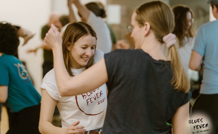 Mehrere Personen tanzen in einem hellen Raum, im Vordergrund lächelt eine junge Frau mit weißem T-Shirt und Schriftzug 'BLUES FEVER 2023'