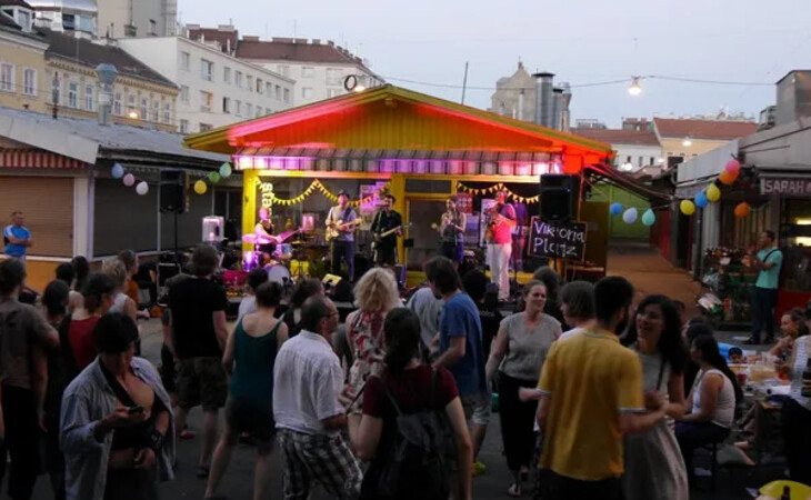 eine Menschenmenge, die nachts eine Straße entlanggeht, beleuchtet von farbigen Lichtern und umgeben von Gebäuden mit Fenstern. Auf der rechten Seite sieht man einen Tisch mit Flaschen und anderen Gegenständen, und im Hintergrund ist der Himmel zu erkennen