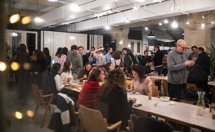 eine große Gruppe von Menschen, die sich in einem großen Raum mit hoher Decke und sichtbaren Rohren versammelt haben. Der Raum wirkt wie ein Coworking-Space, da überall Tische und Stühle verteilt sind. Die Menschen sind unterschiedlichen Alters, Geschlechts und verschiedener ethnischer Herkunft und befinden sich in Gesprächen und im Austausch miteinander. Einige stehen, während andere an den Tischen sitzen. In der Mitte des Raumes steht ein Mann, der zu der Gruppe spricht. Das Bild ist leicht unscharf, wodurch es eine traumartige, ätherische Atmosphäre erhält.