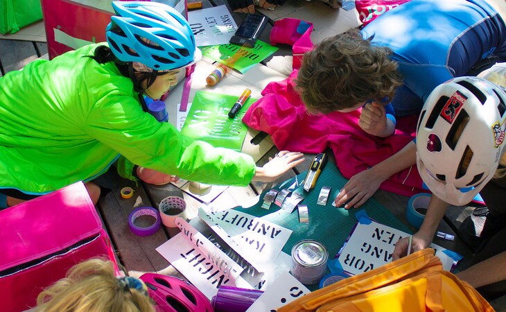 Menschen mit Fahrradhelmen sitzen um einen Tisch und gestalten mit Schablonen und Farbe Plakate
