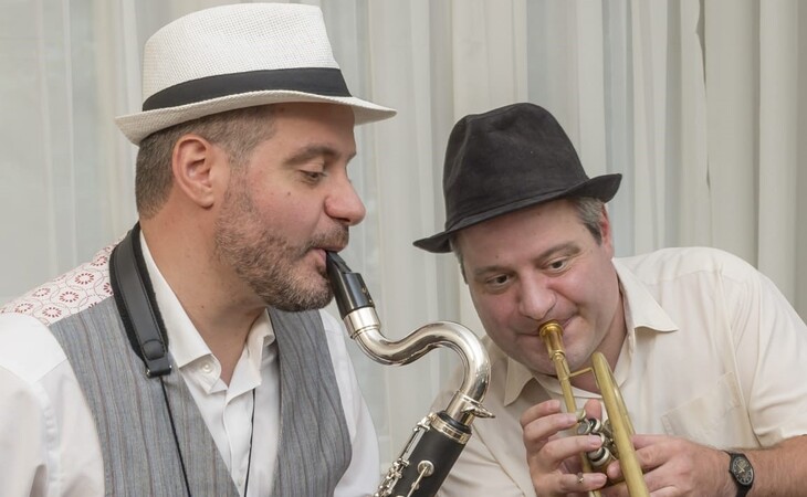 Das Bild zeigt eine Gruppe von Menschen, die vor einer Wand mit Graffiti stehen. Die Frau in der Mitte hält ein Buch in der Hand, während der Mann rechts von ihr eine Tasche trägt. Im Hintergrund sind Fahrzeuge auf der Straße, Gebäude mit Fenstern und ein klarer blauer Himmel zu sehen
