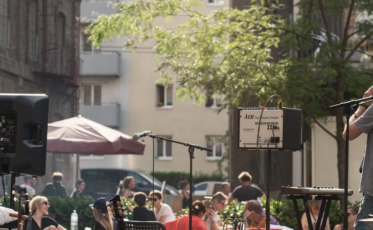 Herbstfest © Julio Arellano Zu sehen ist ein Musiker, der mit seinem Rücken zu uns gewandt ist. Er spielt gerade Mundharmonika. Um ihn stehen Mikrofone und ein Schlagzeug. Im Hintergrund des Bildes sitzen Menschen an Tischen.
