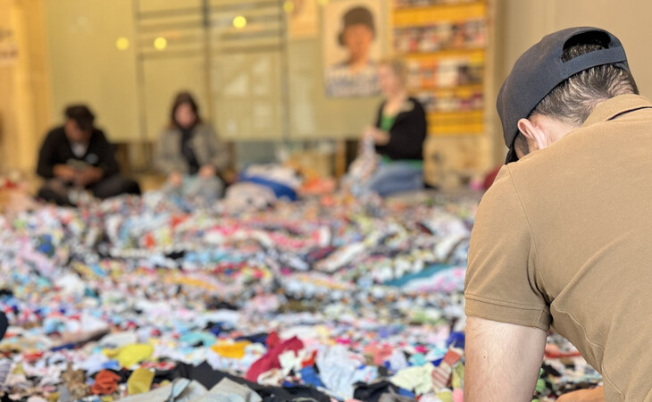 einen großen Haufen weggeworfener Kleidung und anderer Gegenstände, die auf dem Boden eines Raumes verstreut sind. Mehrere Menschen sind im Bild zu sehen; einige sitzen, andere stehen, und sie scheinen die Gegenstände zu sortieren. Die Kleidungsstücke sind unterschiedlich farbig und groß, einige sind gefaltet und aufeinander gestapelt. Im Vordergrund befindet sich ein Mann, der ein braunes T-Shirt und eine Baseballkappe trägt und offenbar an einem Projekt arbeitet. Der Hintergrund ist unscharf, wirkt jedoch hell, da natürliches Licht durch Fenster in den Raum fällt
